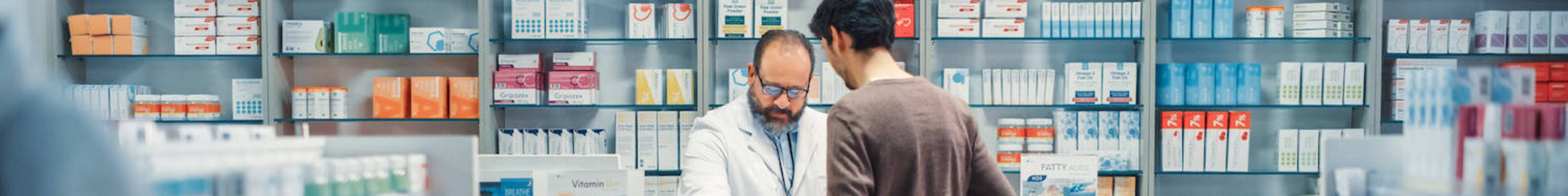 Pharmacy Drugstore Checkout Cashier Counter: Senior Male Pharmacist Scans Barcode and Handsome Young Man Talks to a Cashier and Pays for the Health Care Products at the Checkout Counter.